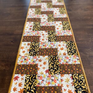 May include: A rectangular quilted table runner featuring a patchwork design of autumn-themed fabrics. The pattern includes various prints of fall leaves, small flowers, and acorns in shades of orange, yellow, brown, black, and cream with a gold border.