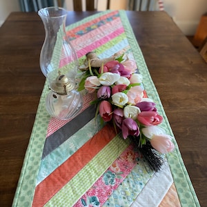 May include: A green, pink, and orange quilted table runner with a chevron pattern. A glass oil lamp and a bouquet of pink and white tulips are on the runner.