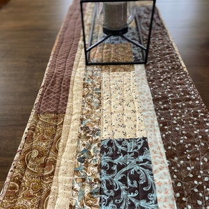 May include: A brown, beige, and blue quilted table runner with a black metal candle holder in the center.