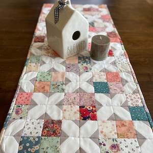 May include: A white quilted table runner with a geometric pattern of squares and diamonds in shades of pink, green, and gray. The runner features a small white ceramic house with the word "Blessed" on the front and a gray candle.