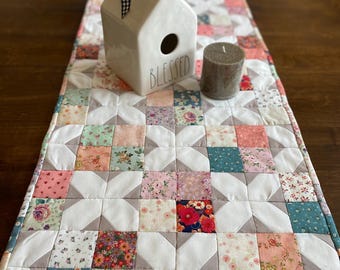 May include: A white quilted table runner with a geometric pattern of squares and diamonds in shades of pink, green, and gray. The runner features a small white ceramic house with the word "Blessed" on the front and a gray candle.