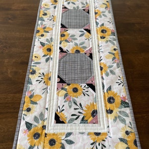 May include: A white table runner with a floral pattern featuring yellow sunflowers, pink flowers, and green leaves. The runner has a white border with a black and white checkered border.