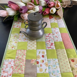 May include: A green and white patchwork table runner with a silver teapot and lid on top. The runner is made of squares of different fabrics with floral and geometric patterns. A bouquet of pink, white, and purple tulips is in the background.