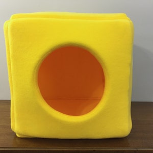 May include: A bright yellow, cube-shaped pet bed with a circular opening for entry. The bed is made of soft, plush fabric.