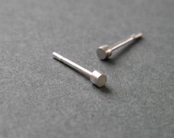 May include: Two silver stud earrings with a round, flat top. The earrings are on a gray surface.