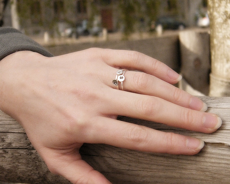 May include: A silver ring with three small charms, each with a letter on it. The ring is worn on a person's finger.