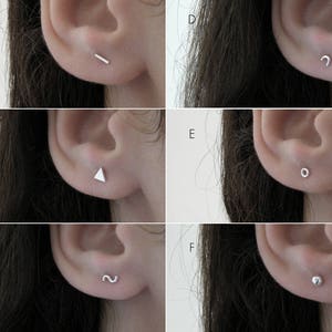 May include: Six close-up images of different silver earrings. The earrings are small and simple, with various shapes, including a line, a triangle, a curved line, a circle, a squiggle, and a small ball.