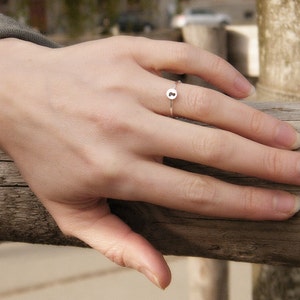 May include: A silver ring with a small round charm featuring the letter "B" on a person's finger.