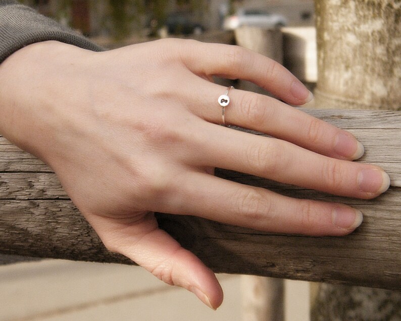 May include: A silver ring with a small round charm featuring the letter "B" on a person's finger.