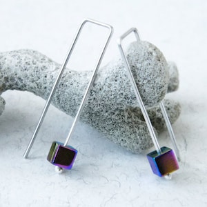 Op de afbeelding: Een paar zilverkleurige oorhangers. Elke oorbel heeft een lange, rechthoekige draad die aan de bovenkant buigt en eindigt met een kleine, iriserende kubus. Aan de basis van elke kubus bevindt zich een kleine zilveren kraal.