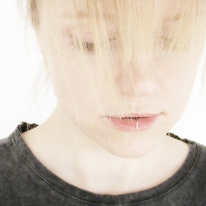 Könnte beinhalten: Eine junge Person mit blonden Haaren schaut nach unten. Ihre Haare bedecken ihre Augen. Sie trägt ein dunkelgraues T-Shirt.