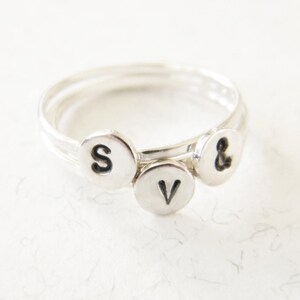 May include: A silver ring with three small charms, each with a letter stamped on it. The charms spell out "S & V".