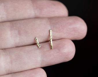 Könnte beinhalten: Zwei kleine, goldfarbene Ohrstecker in Barrenform mit gehämmerten Texturen. Ein Ohrring ist etwas kürzer als der andere. Die Ohrringe werden vor einem neutralen Hintergrund präsentiert, um ihr schlichtes, minimalistisches Design hervorzuheben.