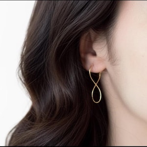 Könnte beinhalten: Nahaufnahme eines Ohrs mit einem goldfarbenen Ohrring in Form eines Unendlichkeitssymbols. Der Ohrring hat ein minimalistisches Design. Die Person hat dunkelbraunes Haar.