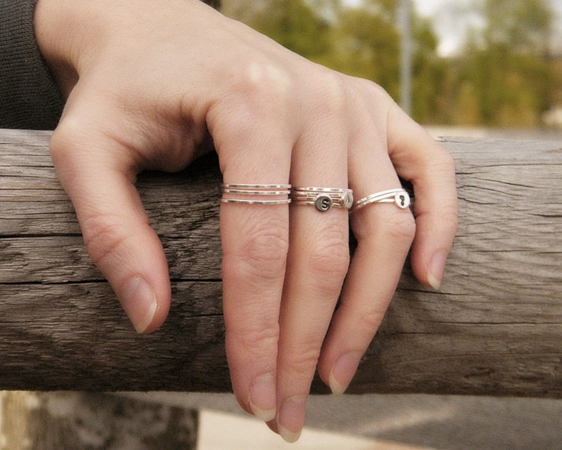 May include: A hand wearing a set of three thin silver stacking rings and a silver ring with a small letter charm.