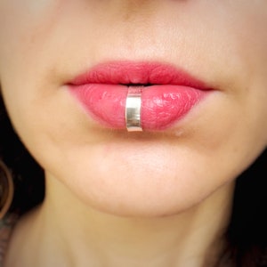 May include: Close-up of a person's lower lip with a silver lip ring. The ring is a simple band with a flat, polished surface.