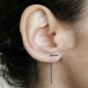 May include: A black bar earring with a long, thin black bar dangling from it. The earring is worn in the upper lobe of the ear.