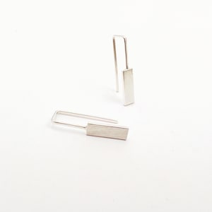 Pode incluir: Um par de brincos de prata com formato retangular. Os brincos têm um design simples e minimalista.