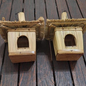 May include: Two handcrafted birdhouses made of natural straw and wood. Each birdhouse features a rectangular body with a rounded entry hole, a thatched roof, and a small wooden perch. The birdhouses are displayed on a dark wooden surface.