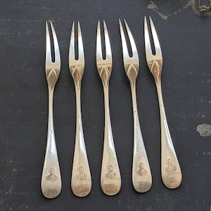 Op de afbeelding: Set van zes zilverkleurige dessertvorken. Elke vork heeft een ontwerp met drie tanden en een decoratief handvat met een gegraveerd monogram. De vorken zijn gerangschikt op een donkere ondergrond, met kant en gedroogde bloemen op de achtergrond.
