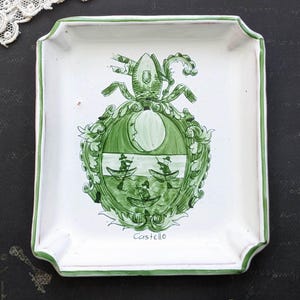 Può includere: Piatto quadrato in ceramica bianca con un emblema decorativo verde. L'emblema presenta un disegno a stemma con una scena di barca e la parola "Castello" in verde. Il piatto ha un bordo verde.