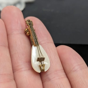 May include: A small, gold-coloured mandolin brooch with a pearlescent body and fine string detail. The instrument's headstock has intricate filigree. A charming, vintage-inspired accessory.