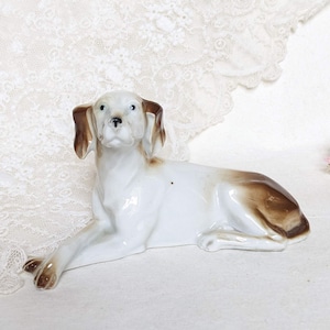 Puede incluir: Figura de perro de cerámica en posición reclinada. El perro es principalmente blanco con marcas marrones en las orejas y la espalda. La figura tiene un acabado brillante y descansa sobre una superficie blanca.