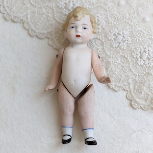 May include: A vintage porcelain doll with blonde hair, blue eyes, and red lips. The doll is wearing black shoes and white socks with blue trim. The doll's arms and legs are jointed. The doll is on a white surface with a lace doily.