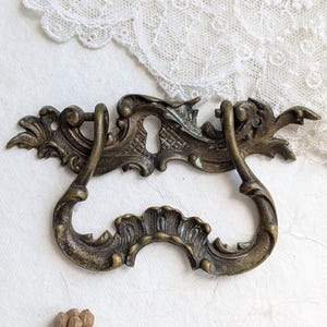 Könnte beinhalten: Antiker Schubladengriff aus Messing mit aufwendigen Details. Der Griff hat ein Schlüsselloch und ein geschwungenes Griffdesign. Die Hardware hat eine Vintage-Ästhetik mit einem verwitterten Finish. Der Hintergrund umfasst eine weiße Spitzendecke und Trockenblumen.