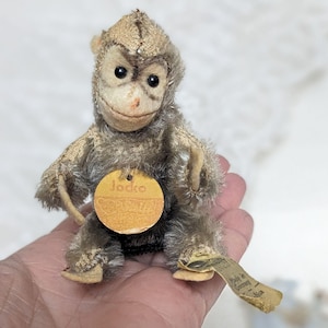 May include: A vintage stuffed monkey toy with a tag that reads "Jocko". The monkey has a light brown body, a tan face, and black eyes. The tag reads "Made in Germany".
