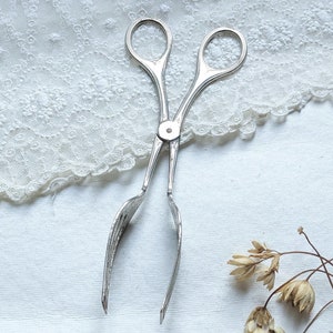 May include: Silver-toned metal tongs with a simple design. The tongs are open and lying on a white surface with a lace overlay.