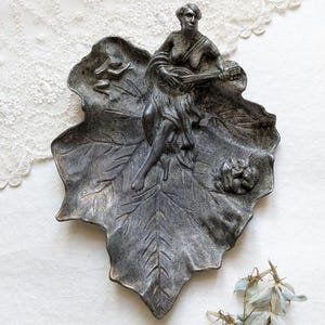 Könnte beinhalten: Eine dekorative Metallschale in Blattform mit einem detaillierten Relief einer Laute spielenden Frau. Die Schale hat eine dunkle, gealterte Oberfläche und aufwendige Blattaderdetails. Die Frau sitzt, und die Schale hat ein klassisches Thema.