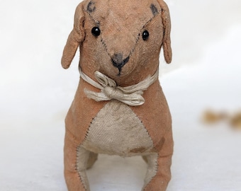 May include: A vintage-style stuffed dog toy with a tan body and head, featuring black button eyes and a stitched nose. The dog has floppy ears and a cream-colored bow tie. The toy is made of a textured fabric.