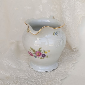 May include: A white porcelain creamer with a gold rim and floral pattern. The creamer has a scalloped edge and a delicate floral design in shades of pink, purple, and blue.