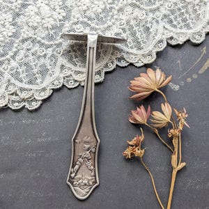 May include: A vintage silver-toned bottle opener with a detailed relief of a person walking with a staff. The opener has a flat, circular top and a long, slender handle. Dried flowers and lace are in the background.
