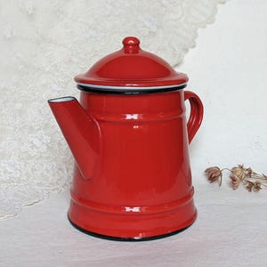 Puede incluir: Una tetera de esmalte rojo brillante con borde y asa negros. La tetera tiene una tapa con un pequeño pomo y un pico vertedor. Esta tetera de estilo vintage es un artículo de cocina clásico.