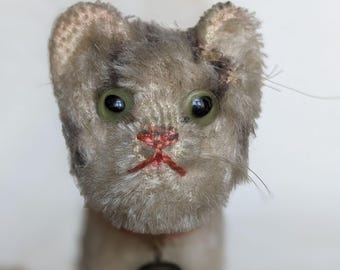 May include: A vintage stuffed cat toy with a gray and white plush body, green eyes, and a red embroidered mouth. It has a small bell on a pink collar. The cat's fur is worn, giving it a charming, antique appearance.