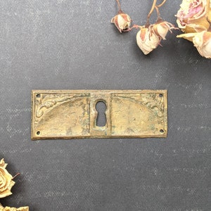 May include: A brass keyhole cover with an ornate design. The cover is rectangular and has a keyhole in the center. The cover is slightly tarnished, giving it an antique look.