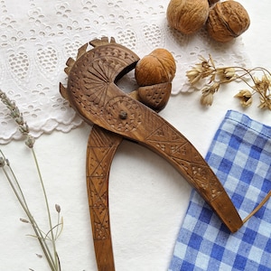 Pode incluir: Um quebra-nozes de madeira vintage com intrincados desenhos esculpidos, segurando uma noz. O quebra-nozes é marrom, com um padrão detalhado na alça e na cabeça. Várias nozes e um pano xadrez azul e branco também são visíveis.