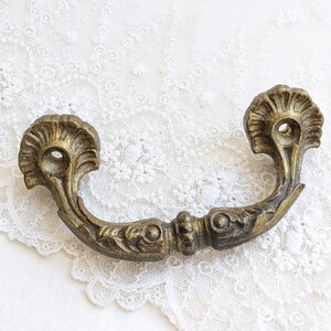 May include: Antique brass drawer pull with a curved handle and ornate floral detailing. The pull features a shell-like design on each end and a central decorative element. The hardware is aged with a warm, golden hue.