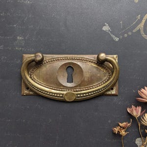Può includere: Maniglia antica in ottone per cassetto con buco della serratura. La maniglia ovale ha un bordo perlato e un manico curvo. L'ottone ha una calda patina invecchiata. Una piccola rosetta rotonda è sotto. Adatta per il restauro di mobili d'epoca.