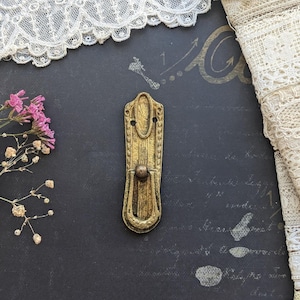 May include: A vertical, elongated brass-toned metal drawer pull with a weathered, textured finish. The hardware features a central drop handle attached to a rectangular backplate with rounded ends and two small circular screw holes at the top.