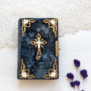 May include: A vintage, rectangular book with a dark blue velvet cover. The cover features a gold cross pendant and ornate gold and white corner accents. A small, gold clasp secures the book. Dried purple flowers are in the background.