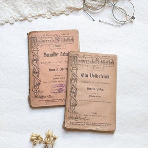 Puede incluir: Dos libros antiguos con cubiertas marrones y letras doradas. Los libros se titulan "Universal-Bibliothek" y presentan el texto "Baumeister Solness" y "Ein Volksfeind".