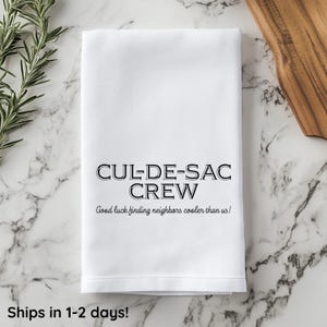 May include: White kitchen towel with black text that reads "CUL-DE-SAC CREW Good luck finding neighbors cooler than us!"