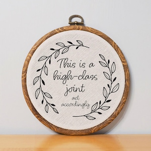 May include: Round wooden embroidery hoop with a cream-colored fabric. The text "This is a high-class joint act accordingly" is centered, surrounded by a black leaf design. A metal clasp is at the top.