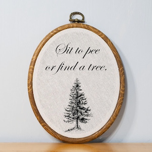 May include: Oval embroidery hoop with a light-colored fabric featuring the text "Sit to pee or find a tree" above a black and white illustration of a pine tree. The wooden hoop has a brass-colored clasp.