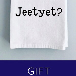 May include: White tea towel with the text "Jeetyet?" printed on it. The towel is folded and tied with twine around a wooden cutting board. The cutting board is 14 inches by 7 inches.  Add-on options include a cutting board and a utensil set.