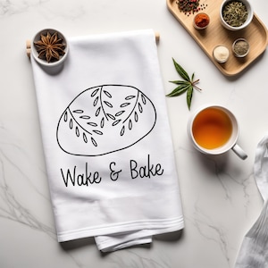 May include: White kitchen towel with a black outline of a loaf of bread and the text "Wake & Bake".