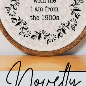 May include: A round, wooden framed wall hanging with a canvas center. The canvas has a floral design and the text "be patient with me I am from the 1900s." The image also includes the text "Novelty Gifts & Unique Home Decor."